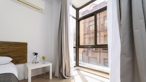 Una habitación de hotel moderna con un ventanal grande, cabecero de madera, mesita de noche blanca con lámpara y jarra, y vista a un edificio de ladrillo.