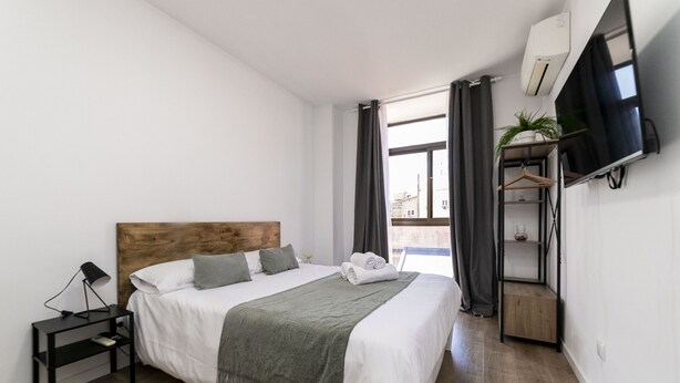 Una habitación de hotel moderna con una cama grande, una mesita de noche con lámpara, un televisor montado en la pared y una planta pequeña en un estante.