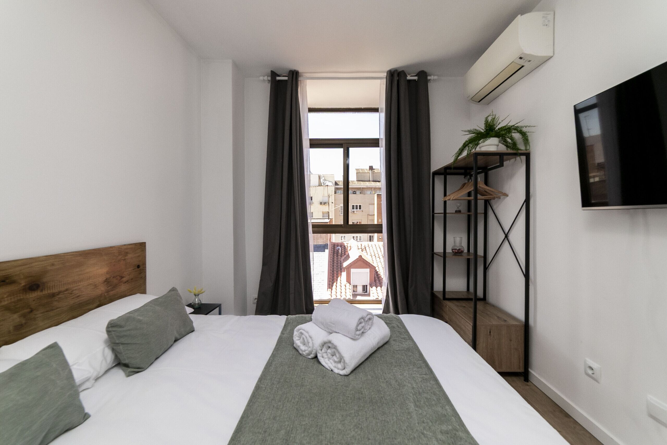 A hotel room with a bed, a TV, a plant, and a view of buildings outside.