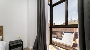 Un dormitorio con un ventanal grande, piso de madera, una mesita de noche negra con un florero y una cama blanca.