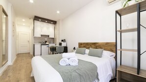 Et moderne hotelværelse med en pænt redt seng, et træhovedgærde, et lille skrivebord med stol, og et tekøkken i baggrunden.
