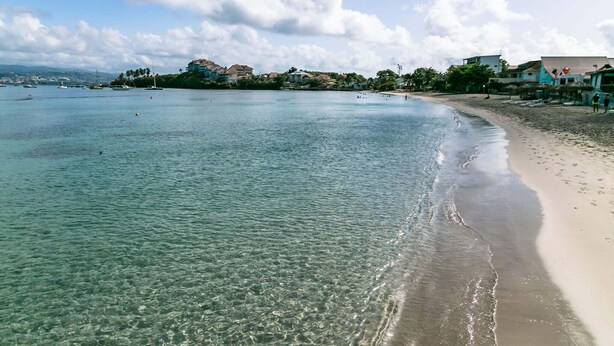 Una playa con agua cristalina, una orilla arenosa y edificios al fondo.