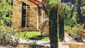 Un bâtiment en pierre avec un toit en tuiles, un grand rocher dans le jardin, ainsi que diverses plantes et arbustes.
