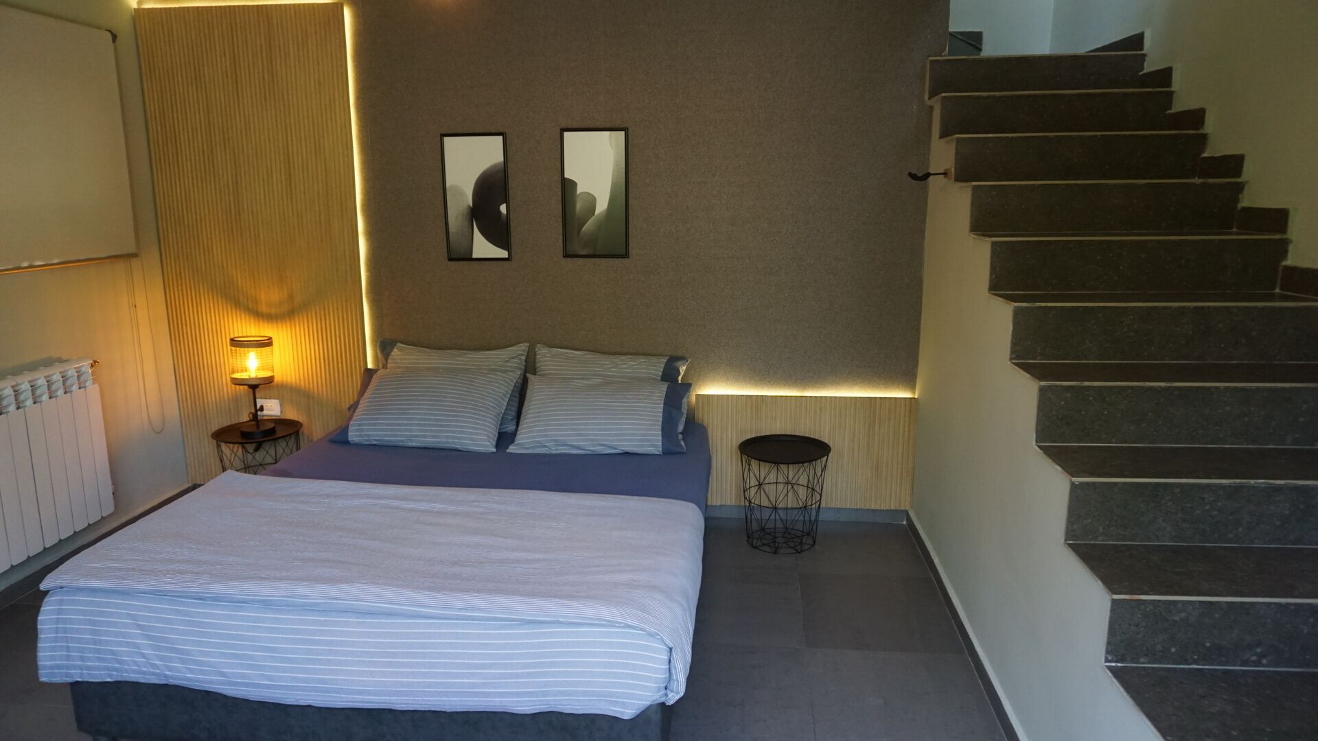 A modern hotel room with a large bed, a bedside table with a lamp, a small round table, and a staircase.