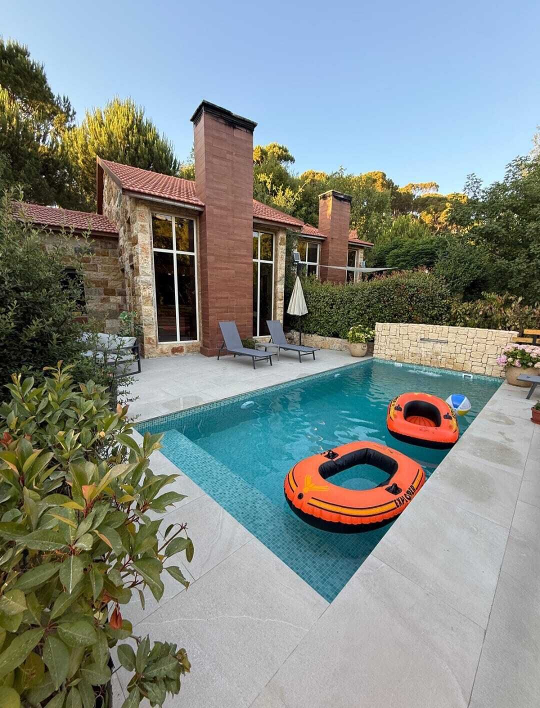 Una piscina con dos flotadores, una pared de piedra y un patio con sillas y una sombrilla.