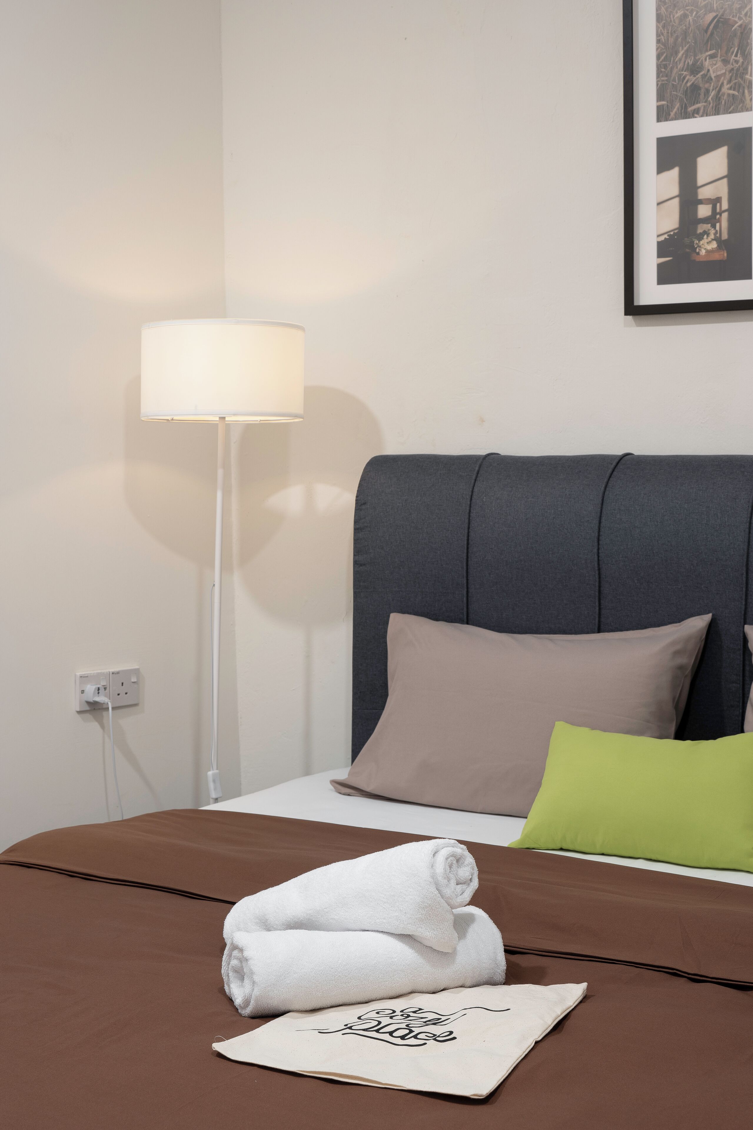 A neatly made bed with a brown bedspread, a rolled white towel, and a small white placemat with text. A floor lamp with a white shade is beside the bed, and a framed picture hangs on the wall.
