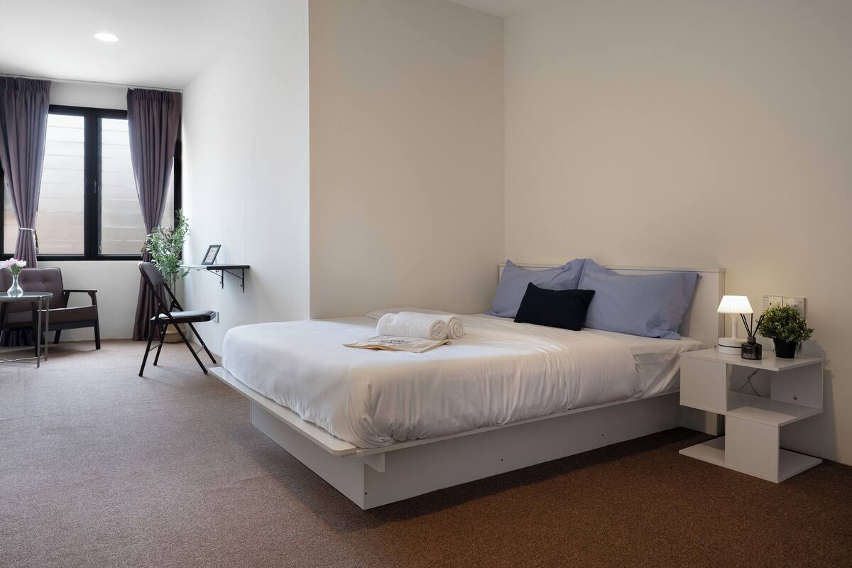 A modern hotel room with a large bed, a desk, a chair, and a nightstand with a lamp and a small plant.