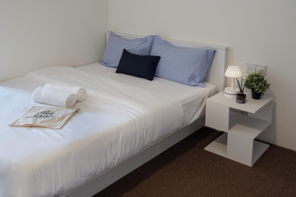 A neatly made bed with white linens and rolled towels, a bedside table with a lamp and a small plant, and a wall socket.