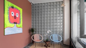 A room with a large abstract painting, two wicker chairs, a small table with a plant, and a view through a glass wall. - A Cozy Place Penang (George Town)