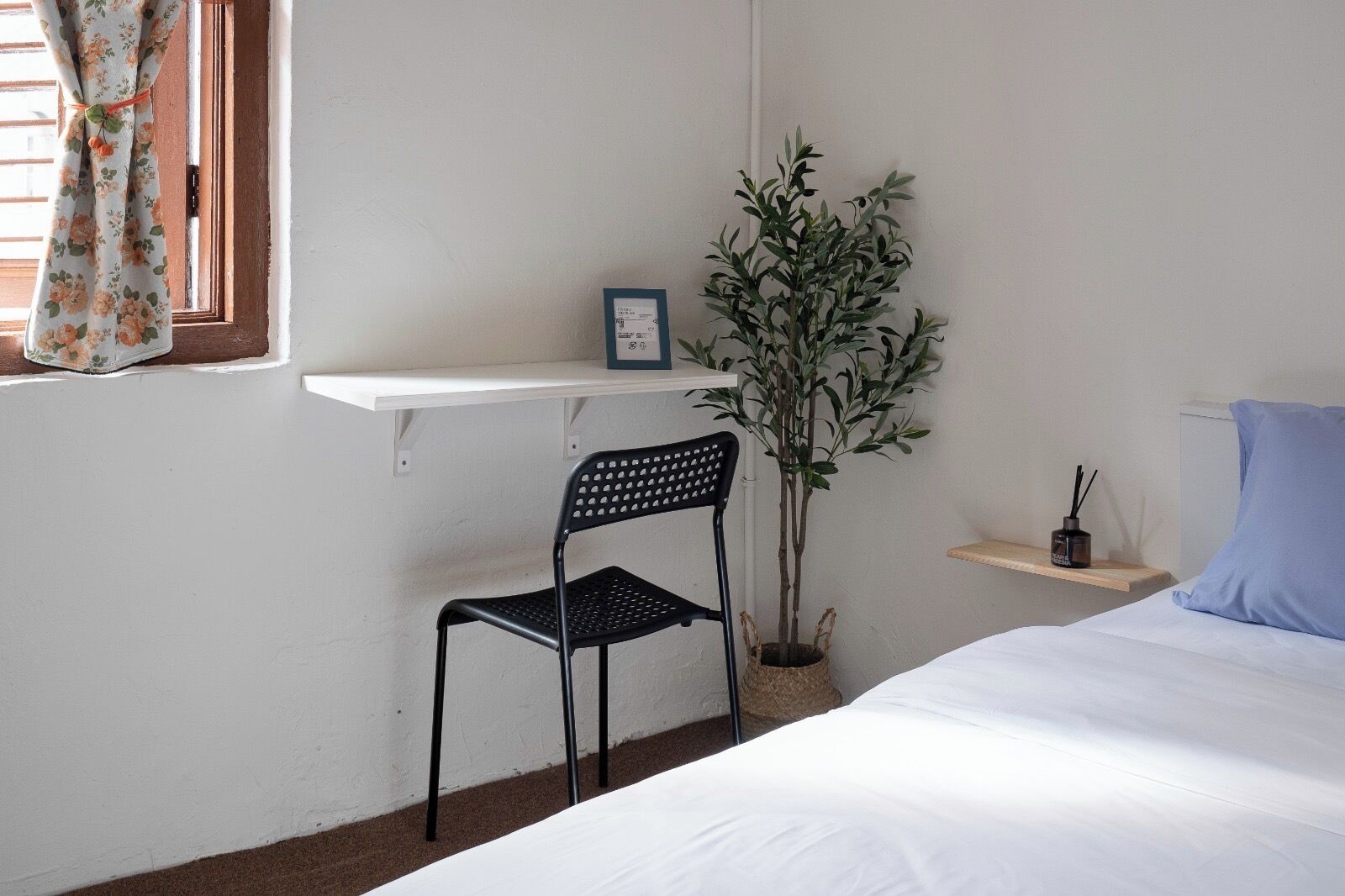 Une chambre à coucher avec un lit, une chaise, une petite étagère sur laquelle est posé un cadre photo, une plante en pot et une fenêtre avec des rideaux.