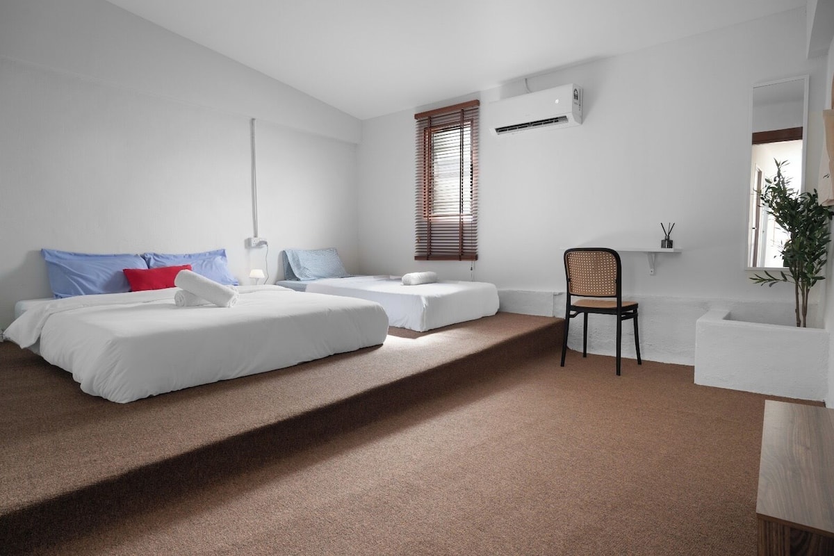 A modern hotel room with two beds, a chair, a small table, and a window with blinds.