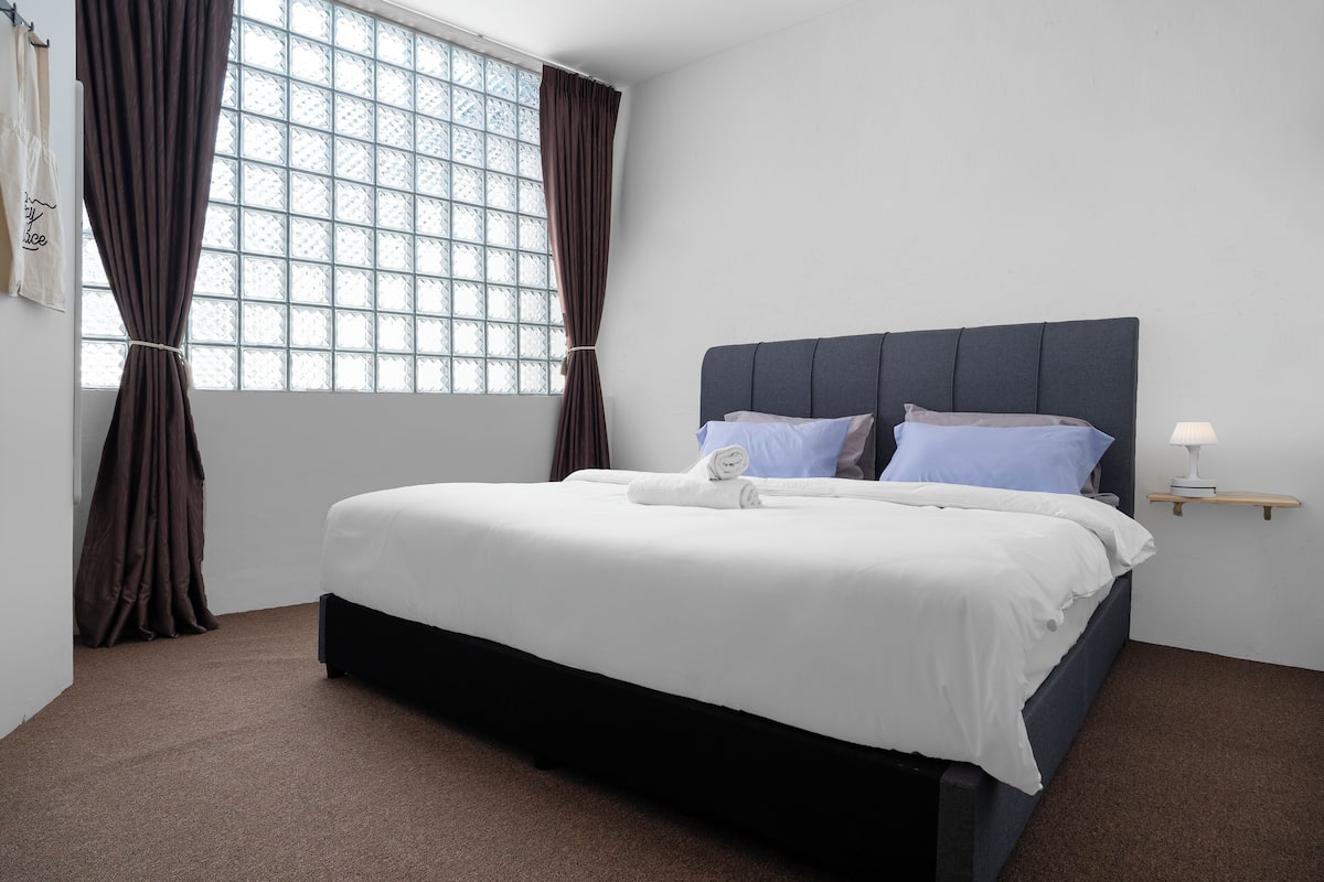 A neatly made bed with white linens and two pillows, a bedside table with a lamp, and a window with brown curtains.