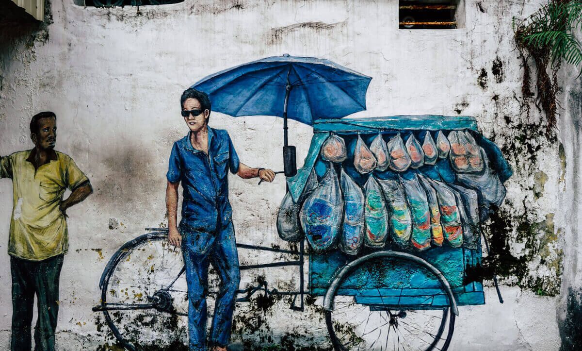 Un mural que muestra a una persona de pie junto a un carrito de bicicleta con bolsas de colores, frente a una pared desgastada.