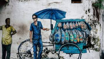 Un mural que muestra a una persona de pie junto a un carrito de bicicleta con bolsas de colores, frente a una pared desgastada.
