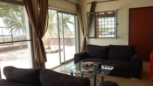 A living room with a dark brown sofa, a glass coffee table, and a view of palm trees through large windows. - Secluded modern villa with pebble beach near Unesco heritage city (Cidade Velha)