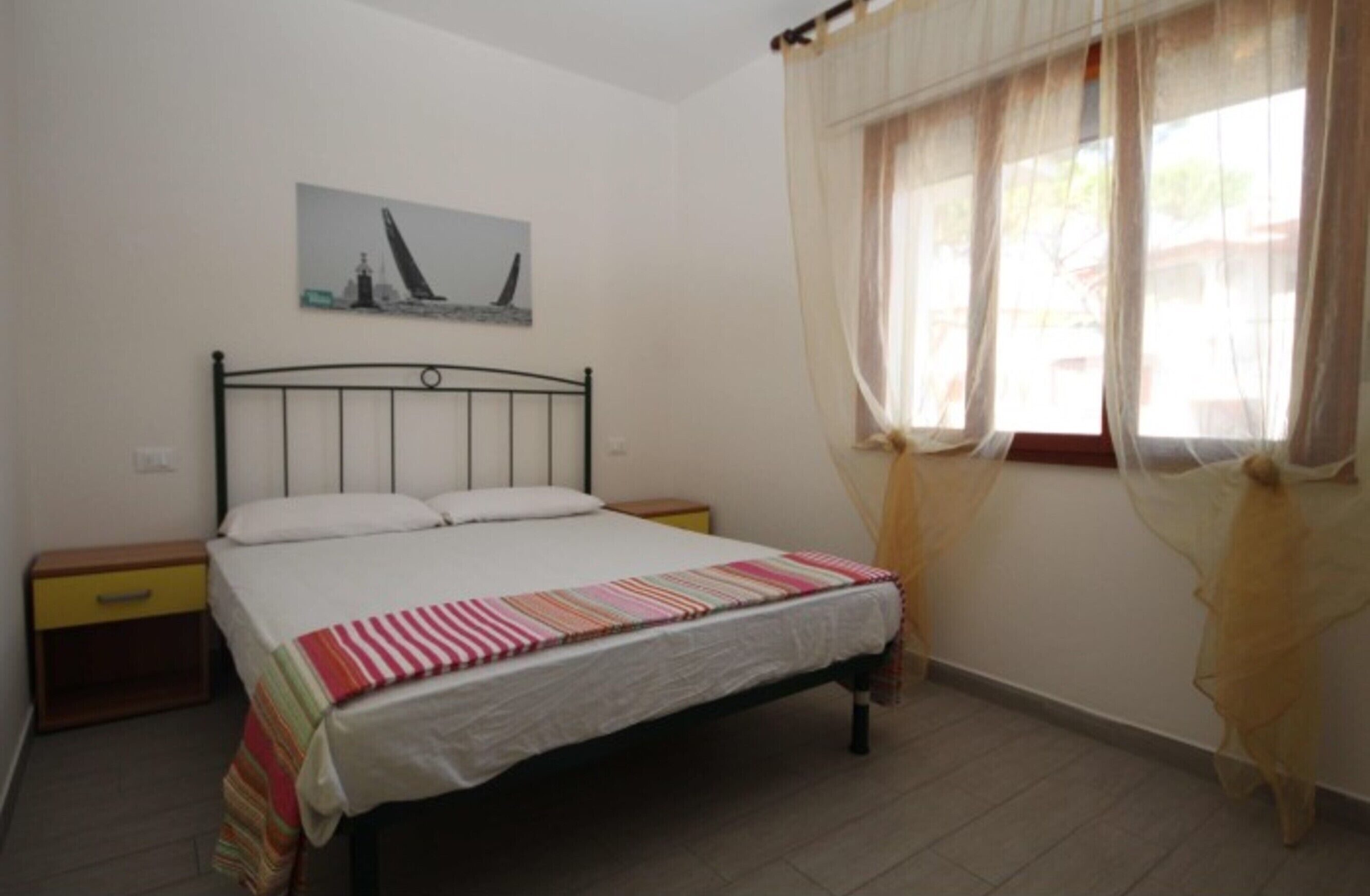 Una cama individual con una colcha a rayas, una mesita de noche, un cajón pequeño y una ventana con cortinas de encaje.
