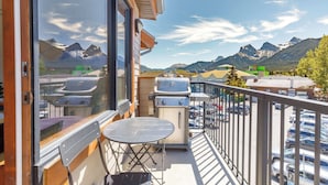 A balcony with a grill, a table, and chairs, overlooking a parking area and mountains.
