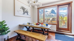 A dining area with a wooden table and bench, a framed animal illustration, and a view of mountains through large windows.