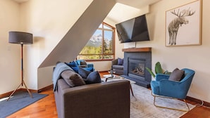 A modern living room with a fireplace, a large TV mounted on the wall, and a mountain view through a skylight.