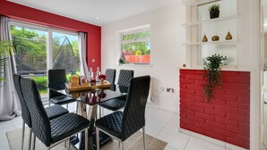A dining area with a glass table and black chairs, a red brick wall, and a window with a view of greenery. -  Elegant 5BR Home with Free EV Charger, Garden and Parking (Slough)