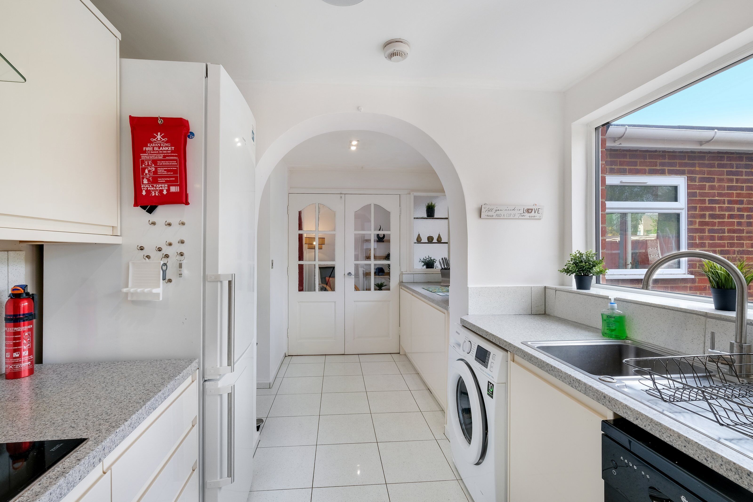 A modern kitchen with white cabinets, a built-in washing machine, a stainless steel sink, and a fire extinguisher.