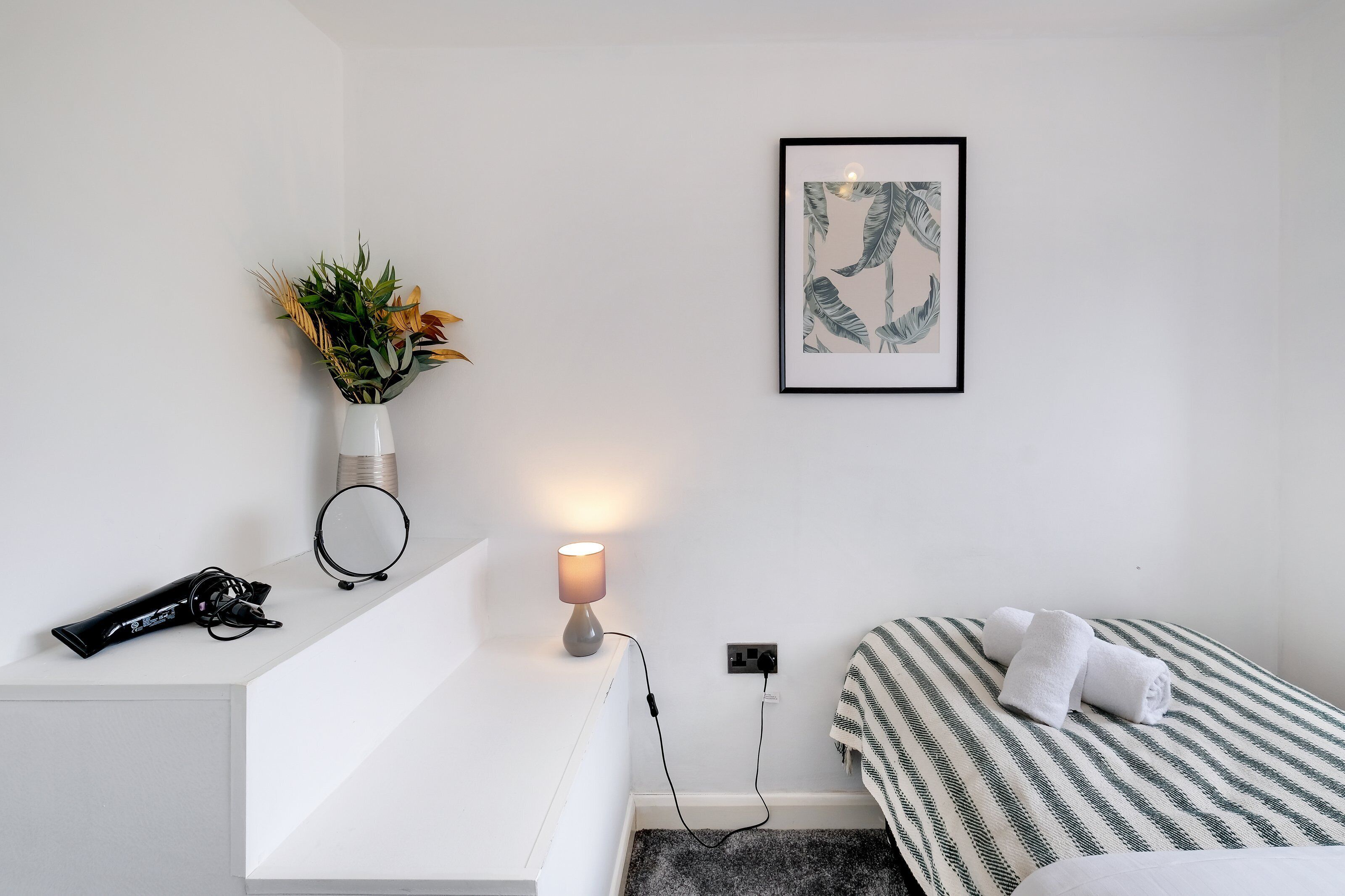 A neatly arranged bedroom with a white bedside table, a lamp, a framed artwork, and a bed with striped bedding.
