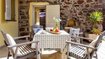 Une terrasse aux murs de pierre, avec une table dressée pour deux personnes, agrémentée d’une nappe à carreaux, de chaises et d’un vase rempli de fleurs.