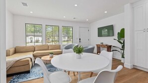 Un salon moderne avec un canapé beige, des chaises blanches, une table ronde et un téléviseur fixé au mur.