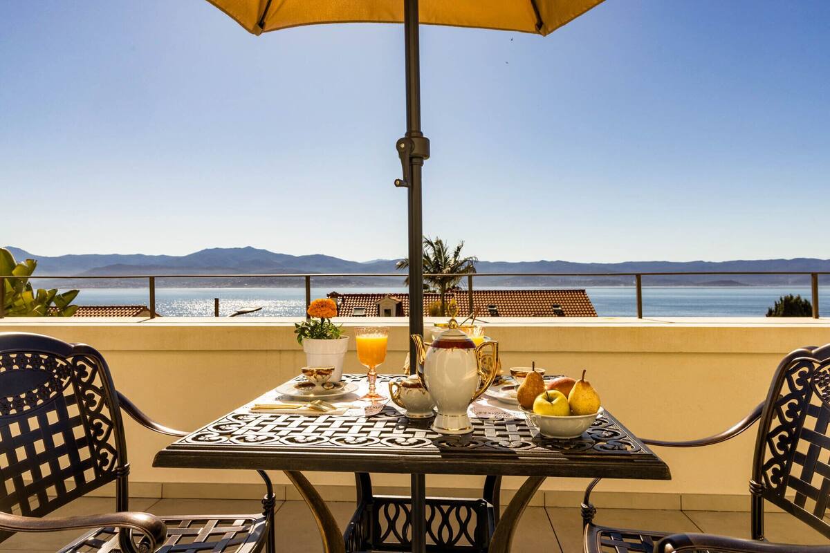 A table with a view of the sea, a yellow umbrella, and a table setting with a pitcher, glasses, and fruit.