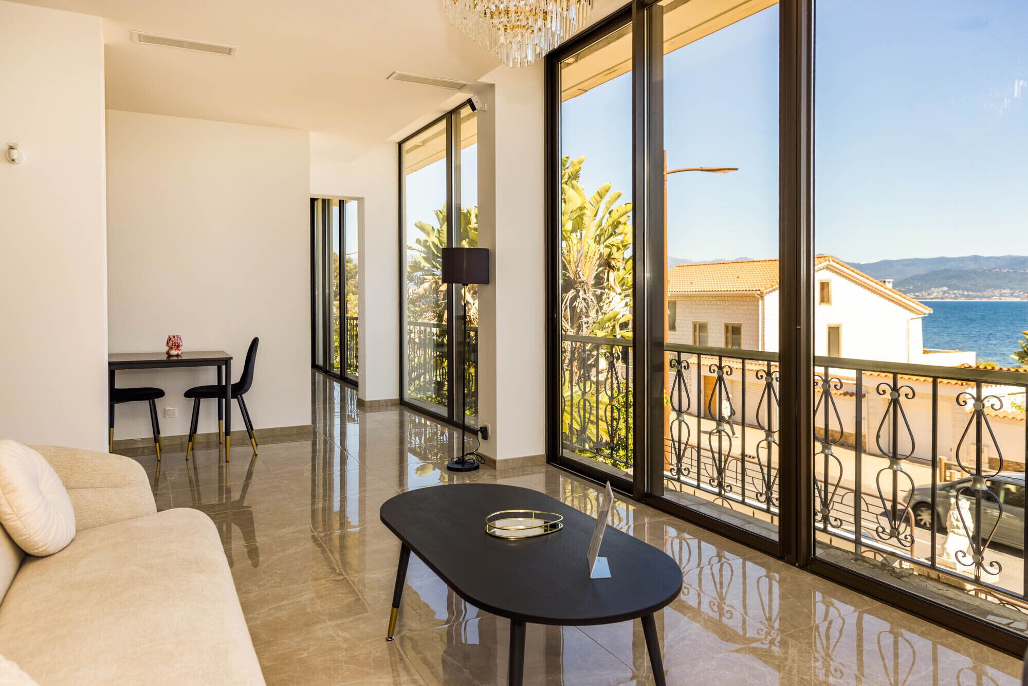 A modern living room with a sofa, a small table, and a balcony with a view of the sea and buildings.