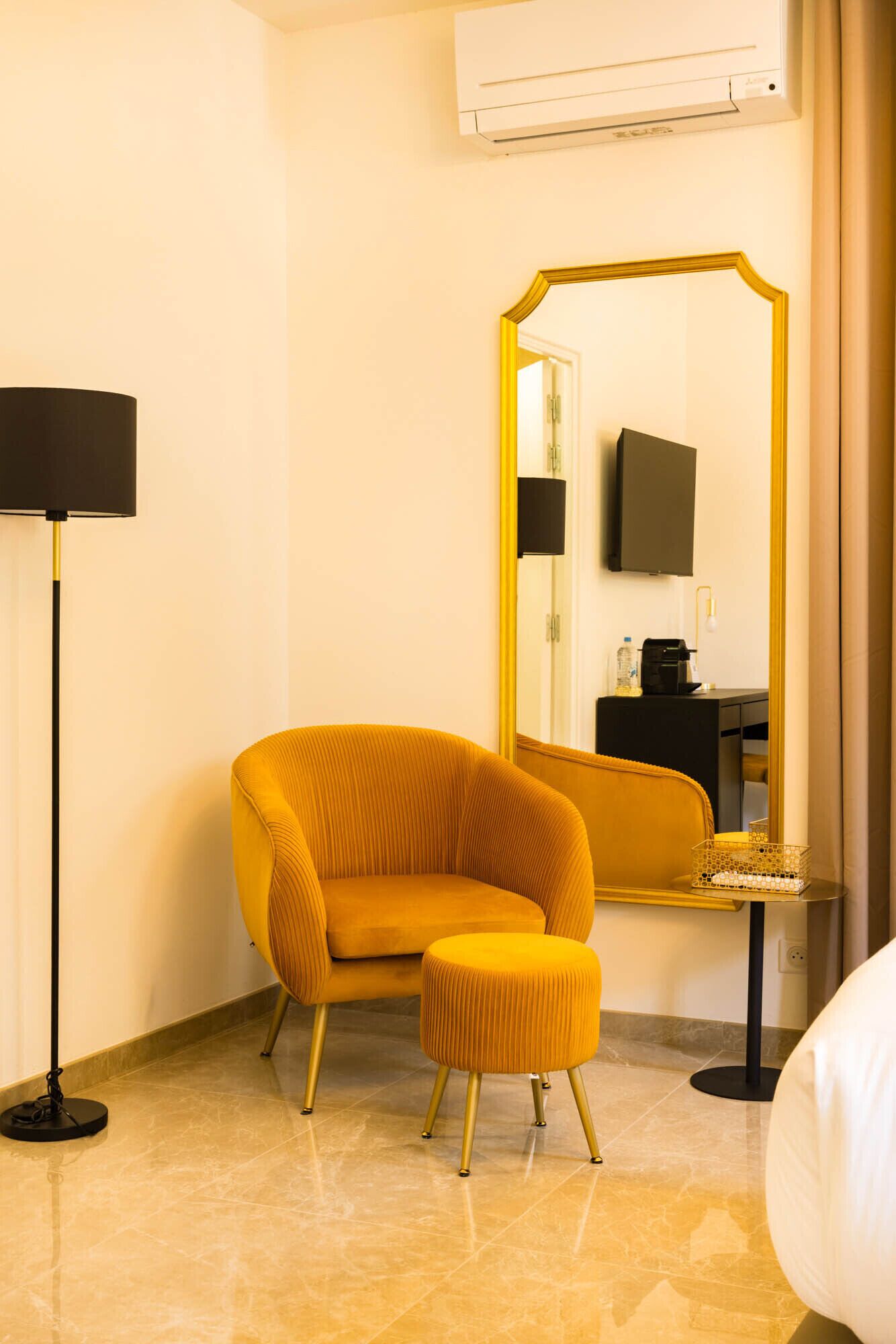 A hotel room with a yellow armchair, a small ottoman, a floor lamp, a mirror, and a flat-screen TV on a black stand.