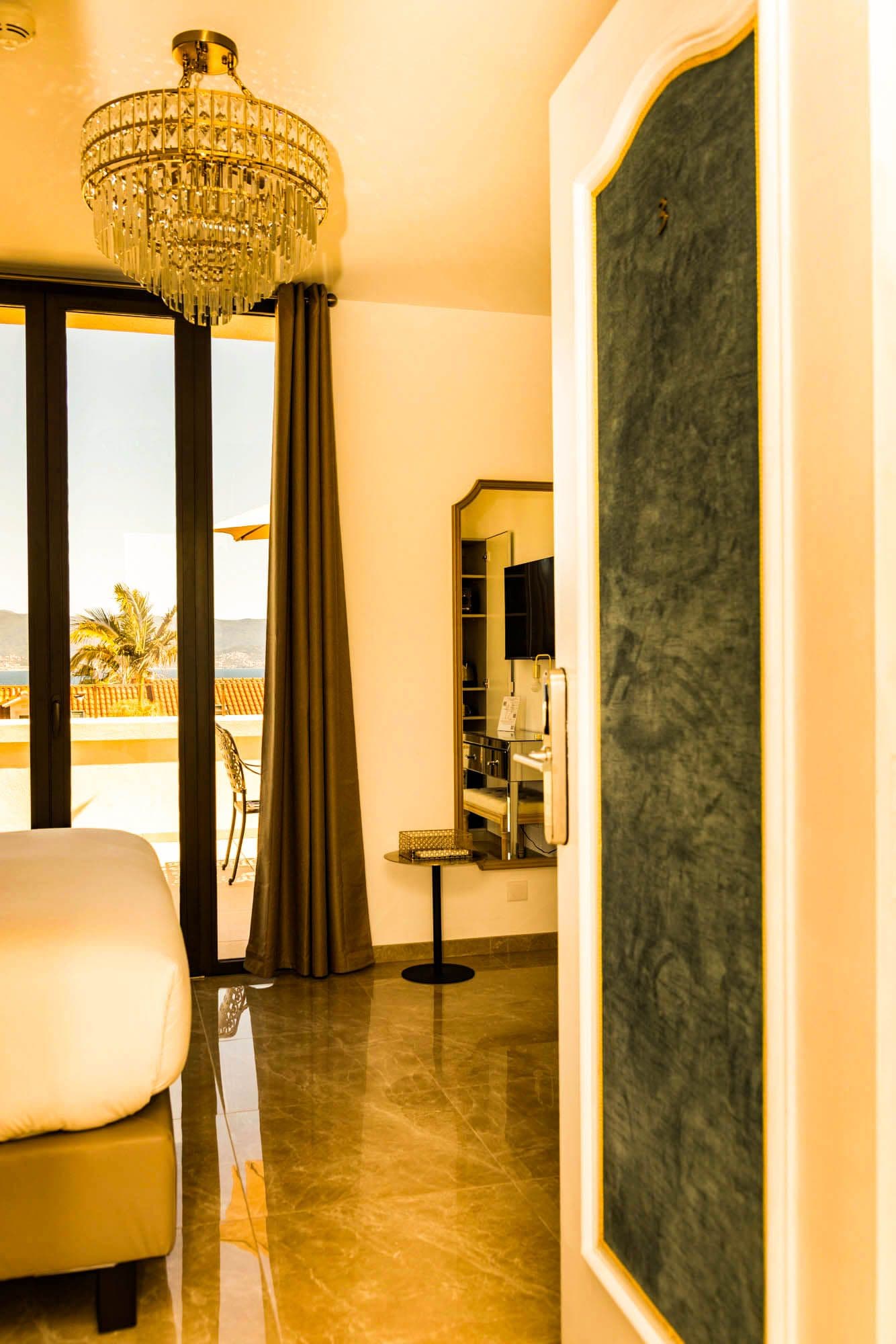 Une chambre d’hôtel avec un grand lit, un lustre, une vue sur la mer et un balcon agrémenté d’un palmier.