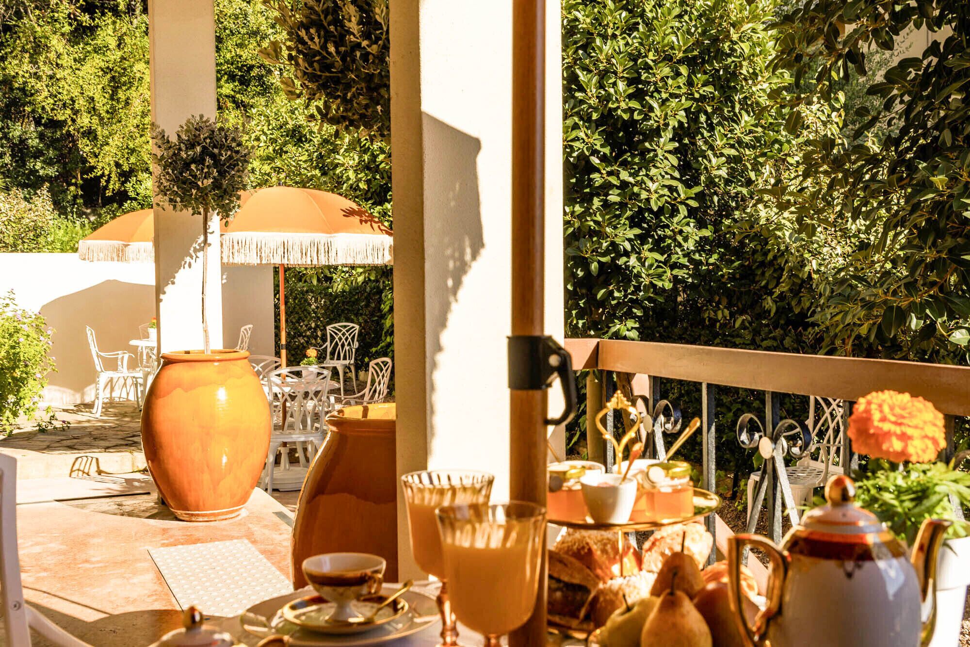 Un espace de restauration en plein air avec une table dressée pour deux personnes, un grand vase et une vue sur un jardin luxuriant.