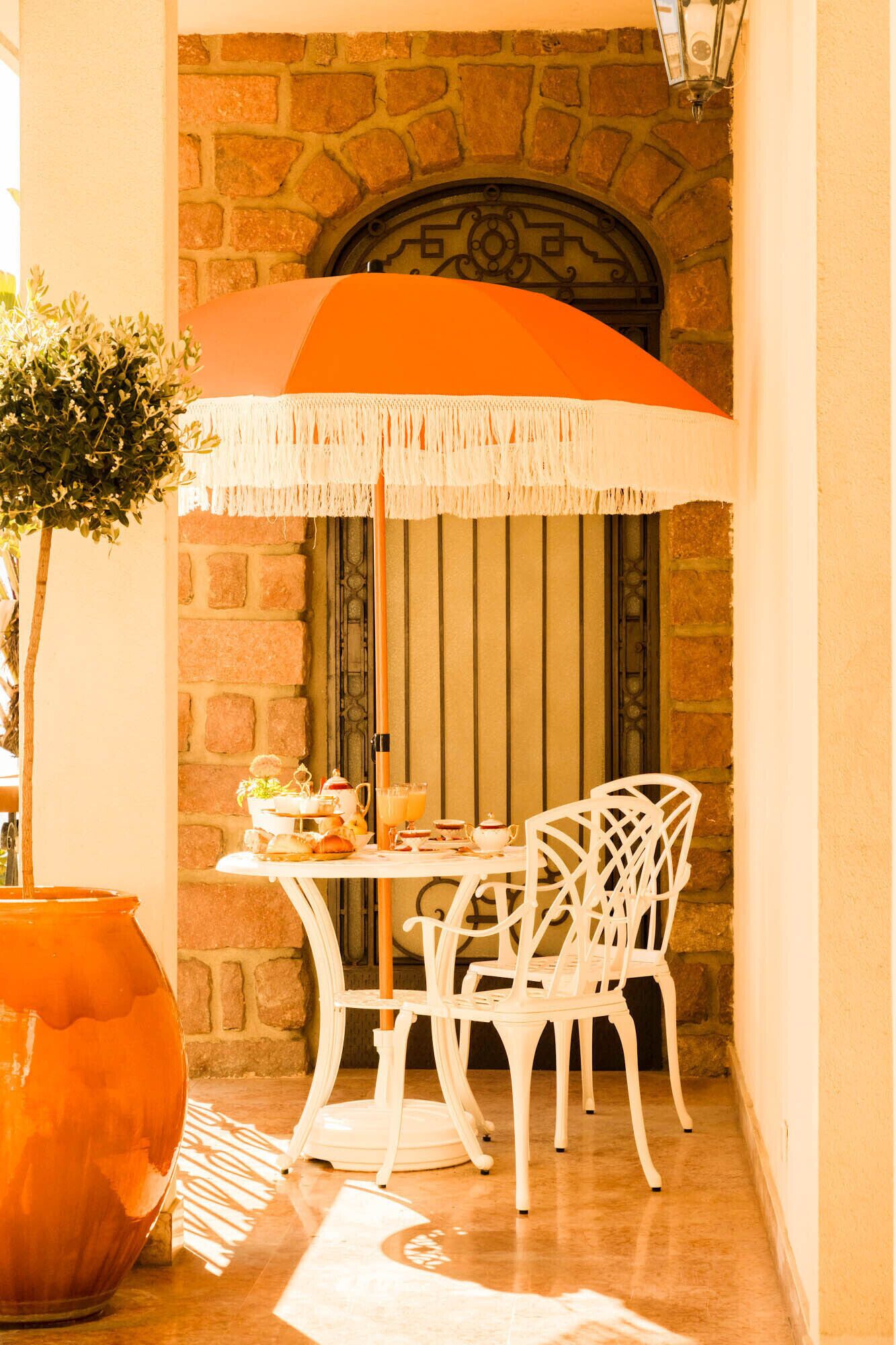 Une terrasse dallée de pierres, avec une table et des chaises, un parasol orange suspendu et une plante en pot.