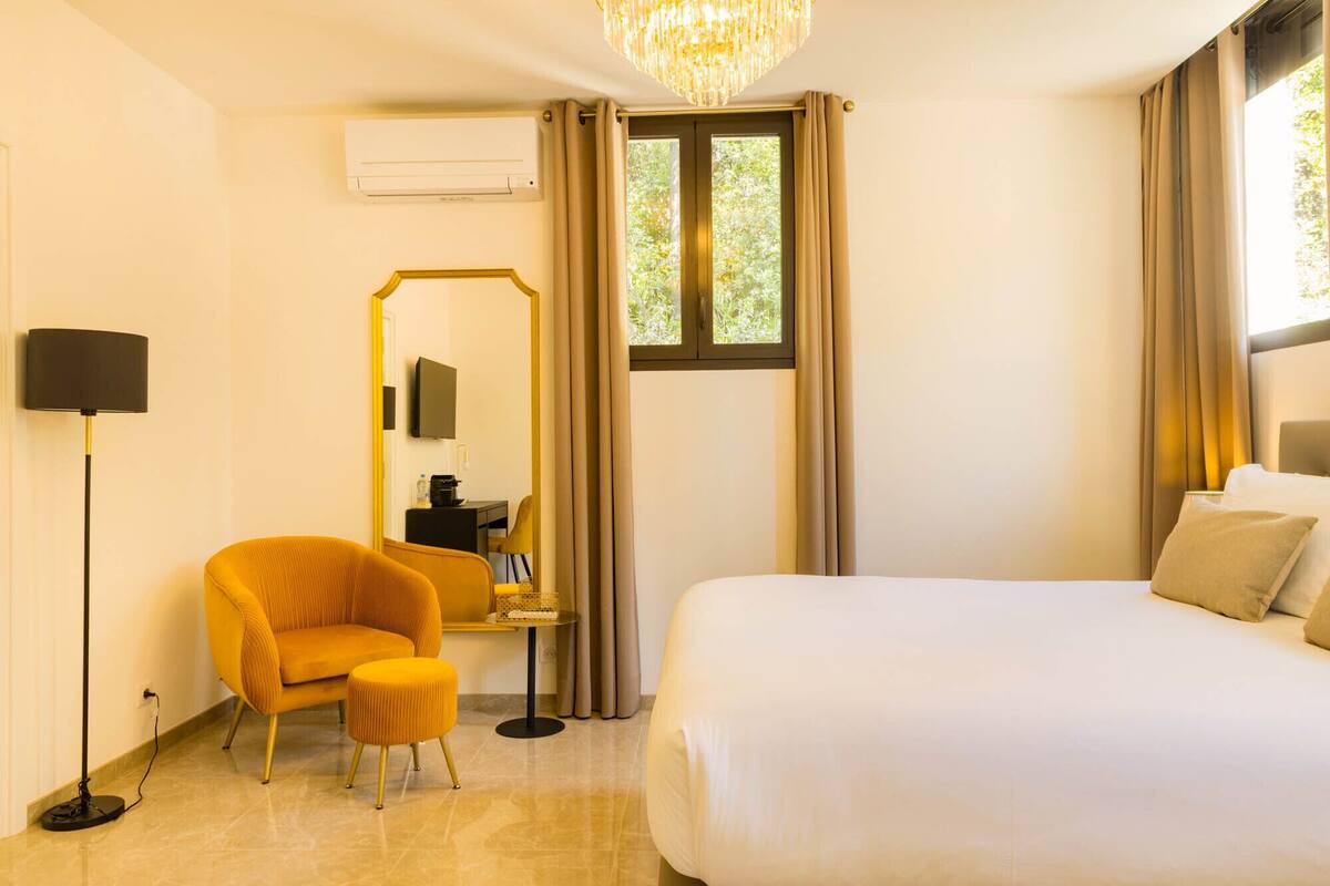 A hotel room with a bed, a chair, a desk with a mirror, a flat-screen TV, and an air conditioning unit.