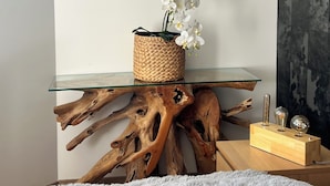 A glass-top table with a woven basket containing white orchids, a wooden block with glassware, and a large piece of driftwood as a base.
