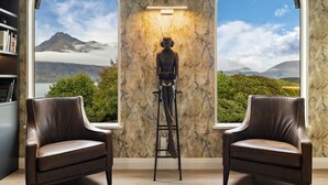 Habitación con vistas a montañas y nubes a través de ventanales arqueados, pared de mármol con una estatua y dos sillones de cuero.