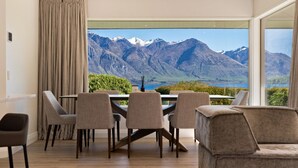 Un comedor con mesa y sillas, vista a montañas y un lago, y un amplio ventanal con cortinas.