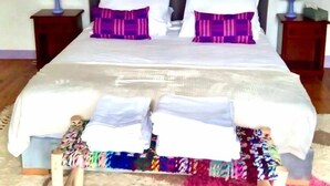 A neatly made bed with white linens and purple pillows, a small wooden bench with folded clothes, and a colorful rug. - LPMDC - A haven of charm and authenticity in the heart of nature (Salerm)