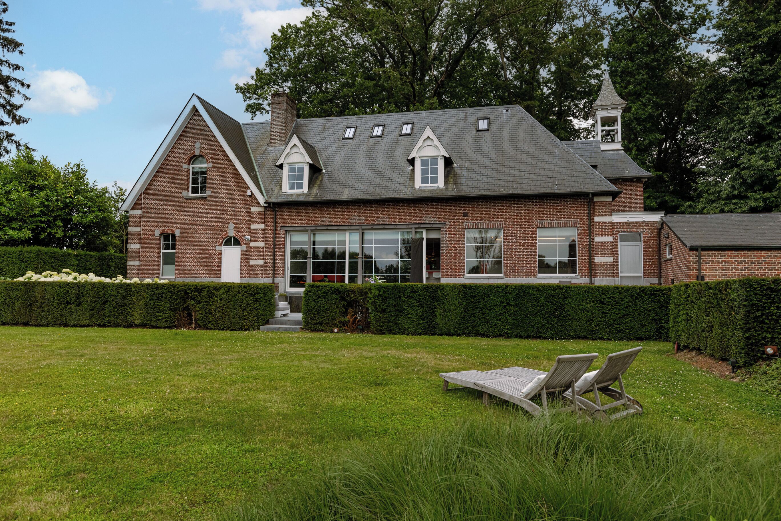 A brick house with a large front lawn, a covered patio, and a tower with a bell.