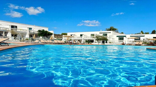 Ein Schwimmbecken mit Liegestühlen und Sonnenschirmen, umgeben von weißen Gebäuden mit Balkonen.