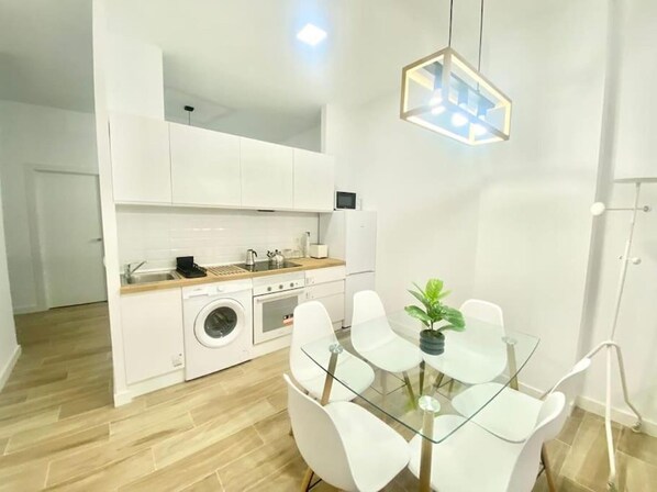 A compact kitchen with a washing machine, a dining table with chairs, and a hanging light fixture. - Prague Bridge Aparment (2-bedroom apartment) (Madrid)