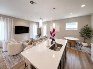 A modern kitchen with a central island, a dining area with a round table, and a living area with a sofa and TV.