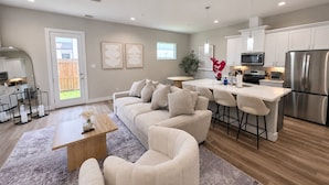 A modern kitchen with stainless steel appliances, a dining area with a white table and bar stools, and a living room with a beige sofa and a coffee table.