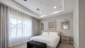 A bedroom with a bed, a bench, a nightstand, a mirror, and a window with curtains.