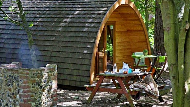 A wooden cabin with a curved entrance, a picnic table with chairs, and a brick wall nearby. - Glamping Pod in Bury St Edmunds Woodland (Bury St Edmunds)