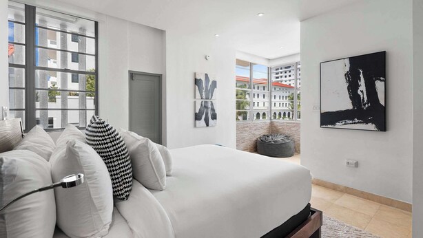 Un dormitorio moderno con una cama grande, ropa de cama blanca y vistas de edificios a través de una ventana.