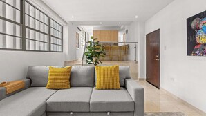 Una sala moderna con un sofá seccional gris, cojines estampados amarillos y una mesita de madera. Una ventana grande con persianas está a la izquierda, y se ve una cocina al fondo.