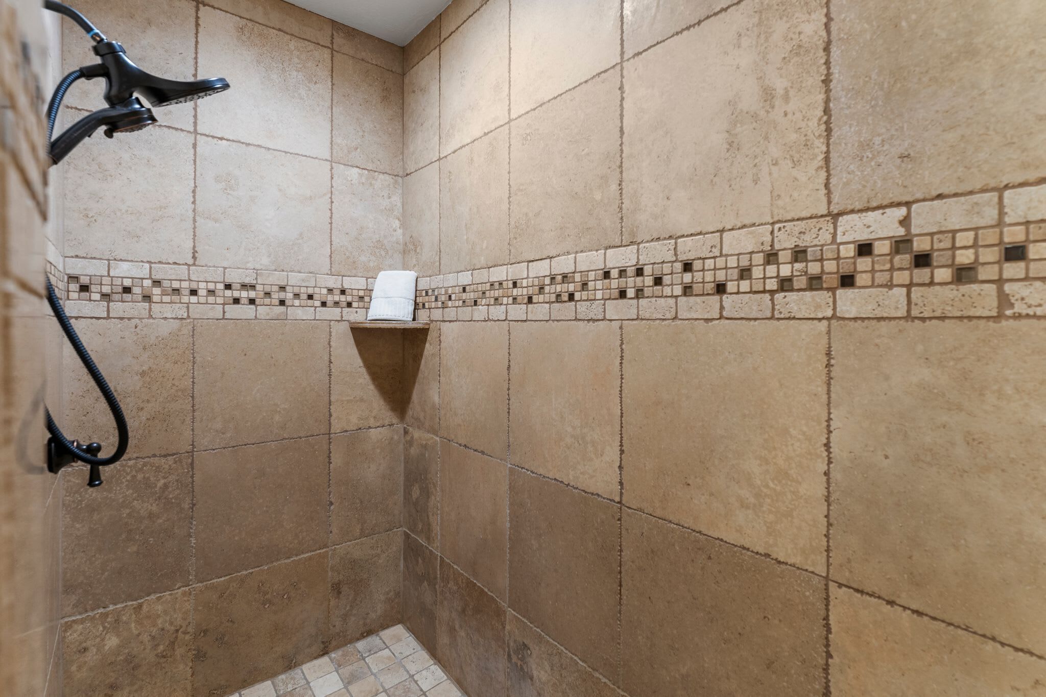 Zona de ducha con azulejos color beige, un estante empotrado y un cabezal de ducha manual.