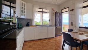 A modern kitchen with white cabinets, a large window with curtains, a dining table with chairs, and a view of the sea. - Villa Branca (Ponta do Sol)